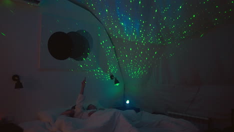 children lie in bed with mother gazing at beams of nightlight on ceiling. young woman points with hand at bright reflections of nightlight in room