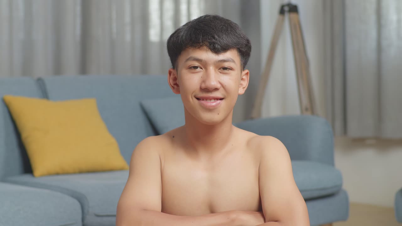 집에서 셔츠를 벗고 운동한 후 덤벨과 함께 매트에 앉아있는 아시아 십대 소년이 팔을 고 카메라에 미소 짓고 있습니다.