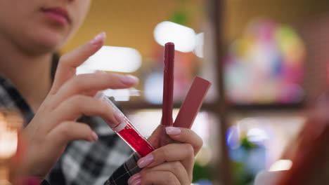 vista de primer plano de una mujer joven examinando e identificando diferentes artículos cosméticos en la mano, incluidos lápiz labial y brillo labial, con luces de fondo borrosas que agregan un ambiente elegante