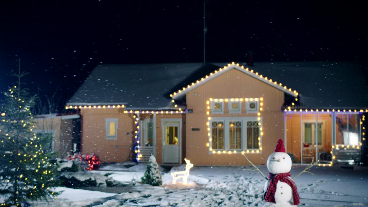 Footage of the Idyllic House Decorated with Garlands on Christmas Eve, Snowman and Christmas Tree stand in the Backyard. Beautiful Winter Evening with Falling Soft Snow.