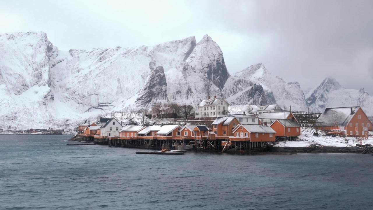 Tradisjonelle oransje rorbuer langs vannkanten i Reine på Lofoten, Norge, under de snødekte toppene av Olstinden og omkringliggende granittfjellrygger, statisk stemningsfullt