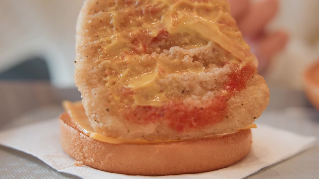 Una vista ravvicinata mostra delle mani che preparano un panino per la colazione su un tovagliolo, posizionando una polpetta di salsiccia rosolata e formaggio fuso su un panino tostato, poi premendo delicatamente sotto una calda luce interna per un pasto fresco e pronto da gustare.