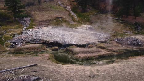 Drone-ellipse-shot-circling-right-of-steam-rising-off-sulfur-natural-hot-spring-boise-national-forest-idaho-located-next-to-a-hillside-and-sulfurous-green-creek-with-coniferous-trees-living-and-dead