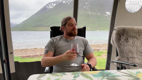 toma estática de un joven con barba bebiendo vino en una carpa durante una fuerte tormenta afuera en verano con una vista panorámica