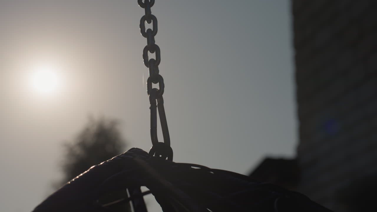 Swing Seat Chain Against Sun Morning Glow, Basket Silhouette And Rooftop Building In Background, Slow Motion Sway And Backlit Hardware, Empty Playground Mood With Urban Nostalgia And Soft Lens Flare
