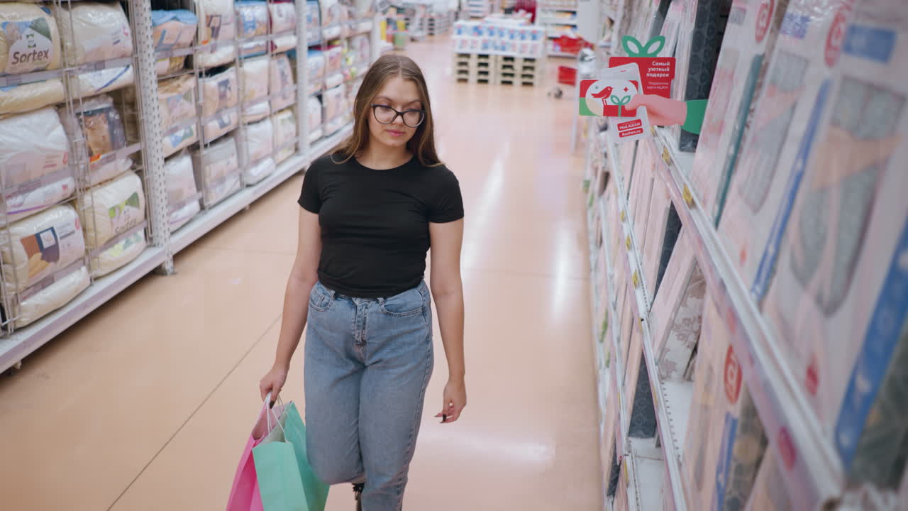 mujer joven con camiseta negra y gafas caminando por el pasillo de la tienda, navegando por las mercancías en los estantes mientras sostiene bolsas de compras, entorno minorista con estantes bien abastecidos y iluminación brillante en el fondo