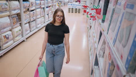 mujer joven con camiseta negra y gafas caminando por el pasillo de la tienda, navegando por las mercancías en los estantes mientras sostiene bolsas de compras, entorno minorista con estantes bien abastecidos y iluminación brillante en el fondo