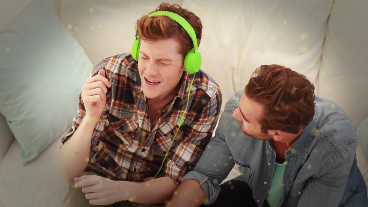 To venner sidder i sofaen og trykker på en telefon for at afspille musik. De nyder musikken i grønne hovedtelefoner og nikker begejstret med hovedet.