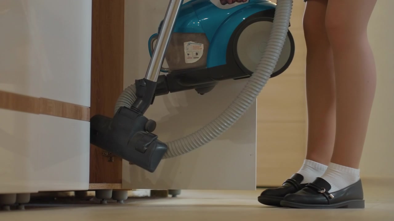 Young white girl finishing kitchen vacuuming, house cleaning complete, legs near clean hardwood floor, vacuum parked beside cabinet, blue canister visible, steady stance, morning light, calm domestic