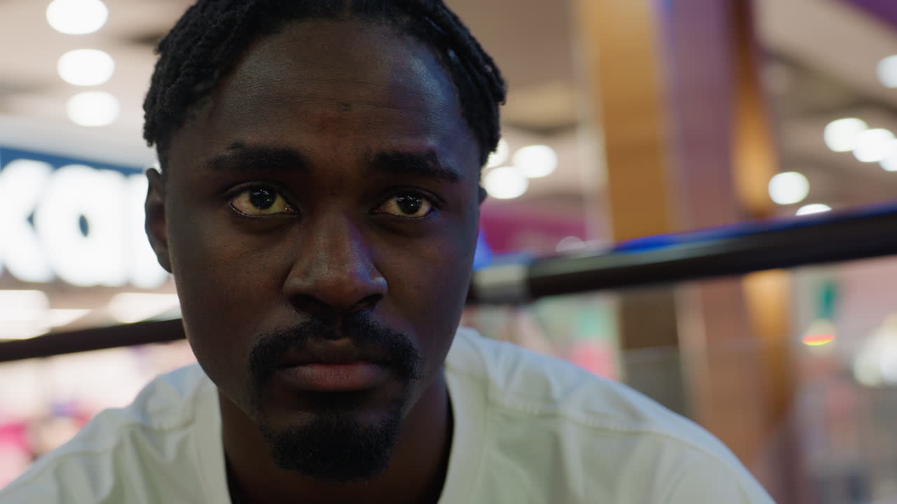 Close up of man with focused expression looking in anticipation seated indoors mall environment as he checks his time with blurred bokeh background creating mood of waiting and curiosity
