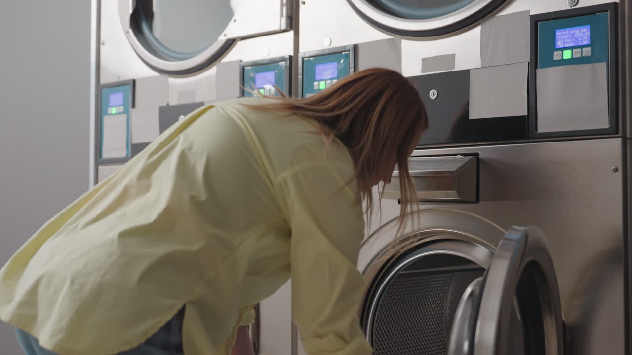 Back view cleaner woman carrying laundry basket through dim laundromat, industrial washers running with fabrics visible behind glass, stainless panels, routine operation, indoor workplace