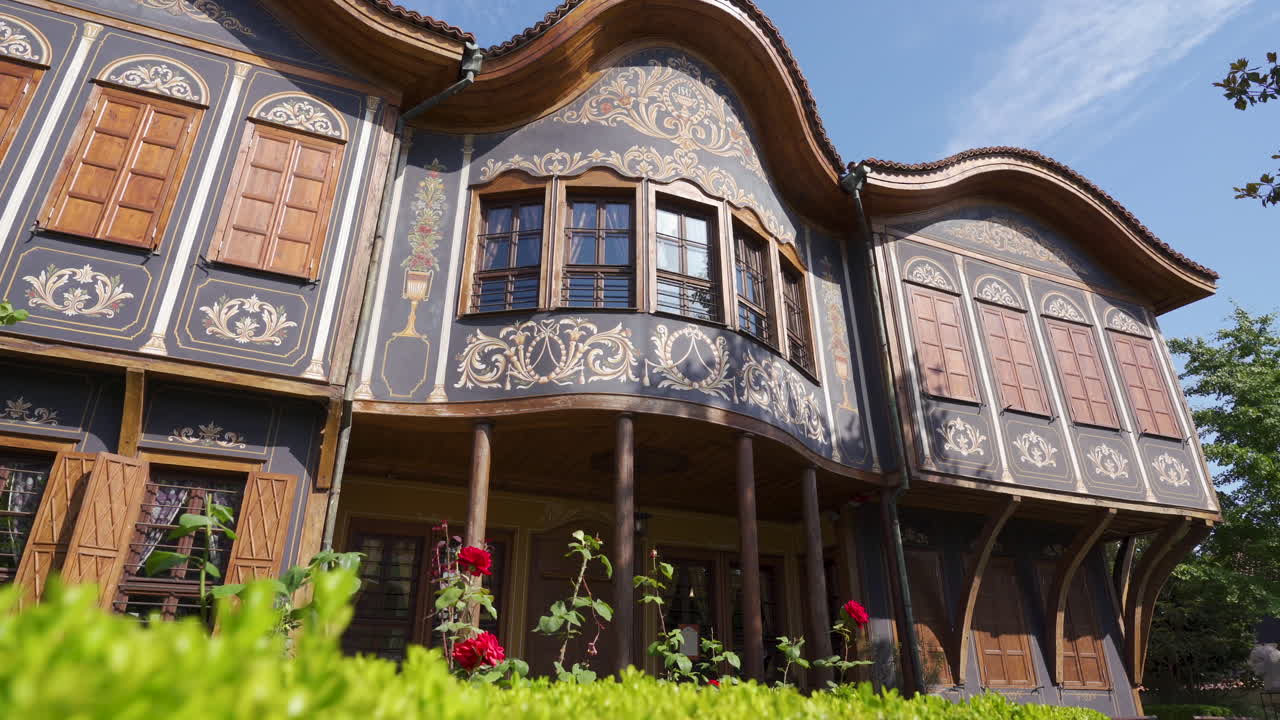 POV shot of the Ethnographic Museum, a richly decorated Revival house with ornate wooden windows, vibrant garden and blue sky backdrop