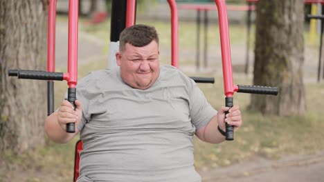a fat young man in a gray tshirt is doing exercises on an exercise machine outdoors a fat young man in a gray tshirt is doing exercises on an exercise machine outdoors struggle with excess weight