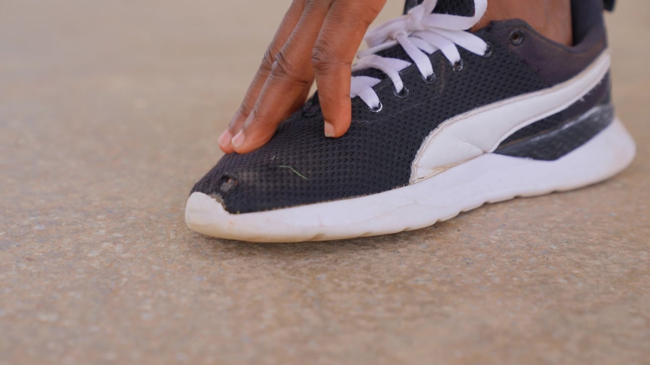 Scarpa sportiva nera usurata con suole bianche su cemento grezzo che mostra danni visibili e interazione manuale, tessuto a rete danneggiato, lacci e usura della calzatura