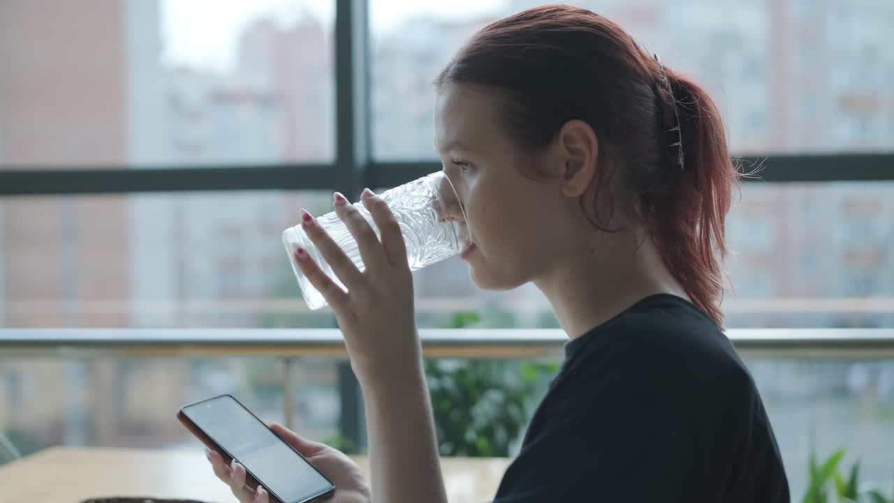 젊은 여성이 큰 창문, 자연빛, 도시 전망을 가진 현대적인 레스토랑에서 나무 테이블에서 신선한 러드를 먹고 있습니다. 편안한 식사 장면