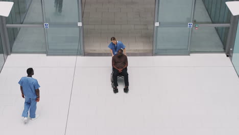 Overhead-View-Of-Nurse-Wheeling-Patient-In-Wheelchair-Through-Lobby-Of-Modern-Hospital-Building