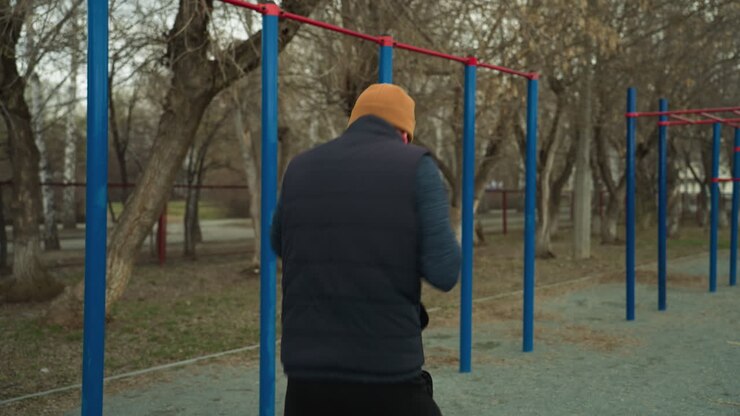 Back view of a coach working out fast in an outdoor gym, with blurred views of a car passing across the road, with red and blue iron bars visible in the background