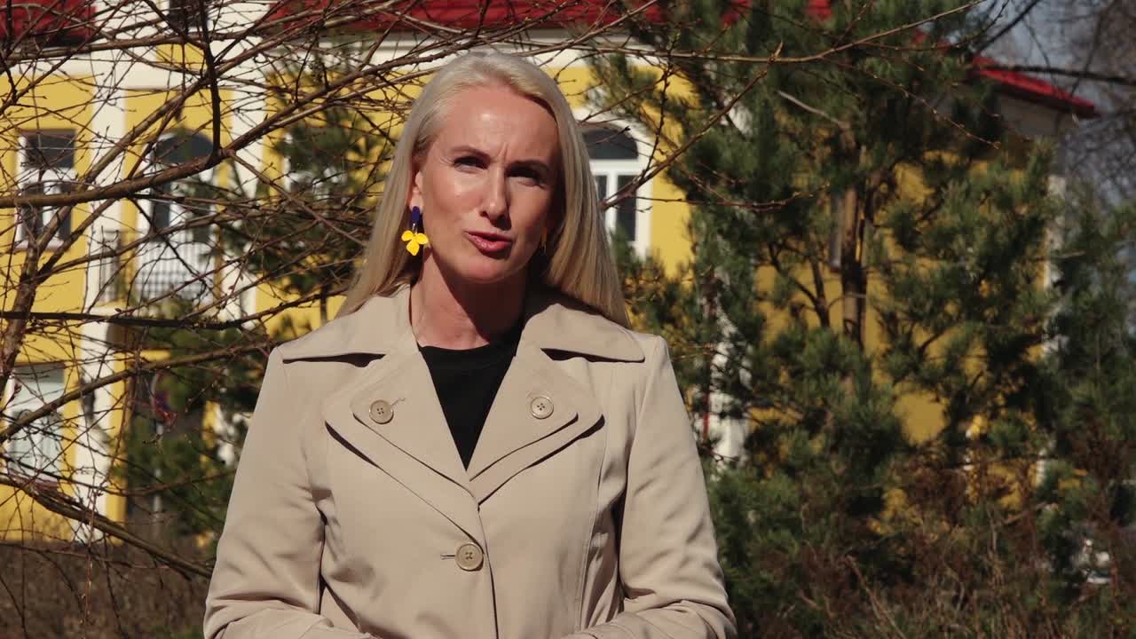 A woman with long blonde hair and colorful earrings smiles confidently while standing outdoors in a beige coat, with trees and buildings in the background.