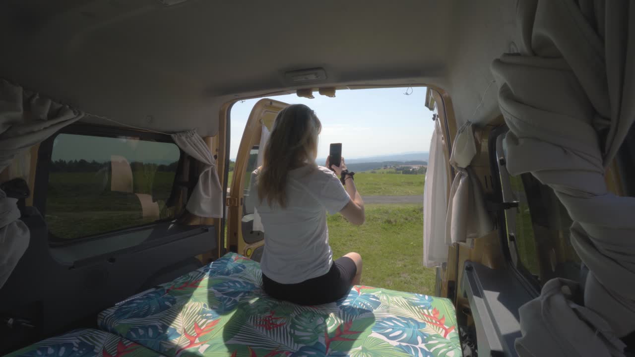 chica nómada digital rubia tomando fotos con el teléfono del paisaje desde la parte trasera de una autocaravana