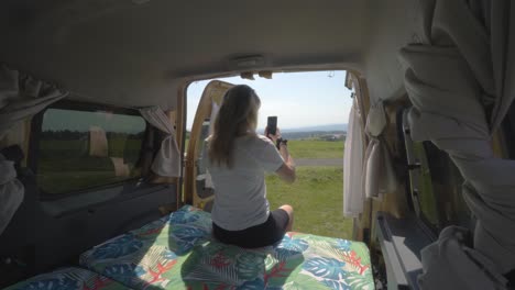 blonde digital nomad girl taking pictures with phone of the landscape from the back of a camper van