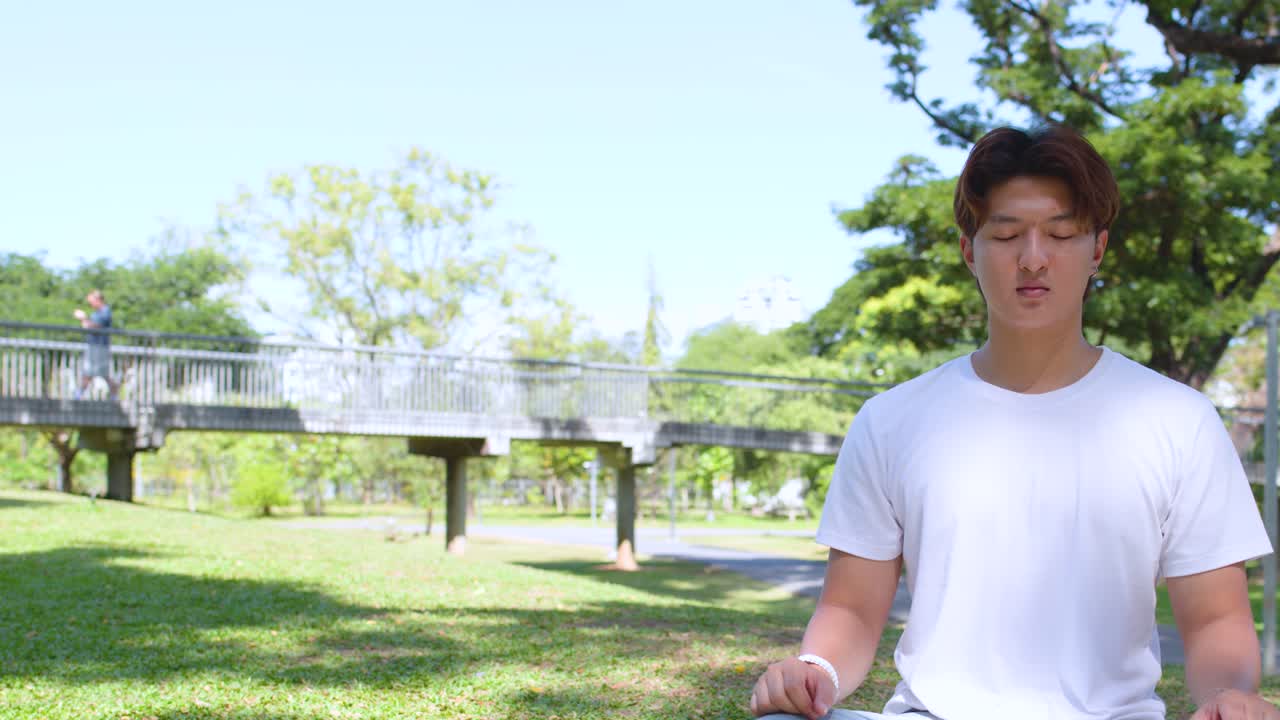 若いアジア人男性が、明るい緑の公共公園に座って、マインドフルネス瞑想を実践しています。