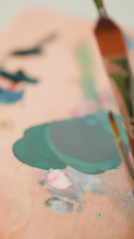 an artist s hand is seen holding a paint palette while delicately mixing paint colors with a brush. the close-up shot captures the careful blending of hues on the wooden palette