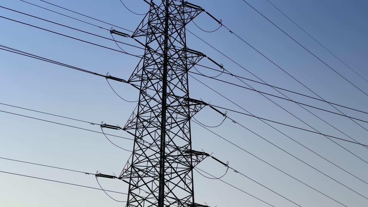 Camera tilts down from the clear sky to reveal a tall steel transmission tower, power lines extending outward against a smooth blue gradient background