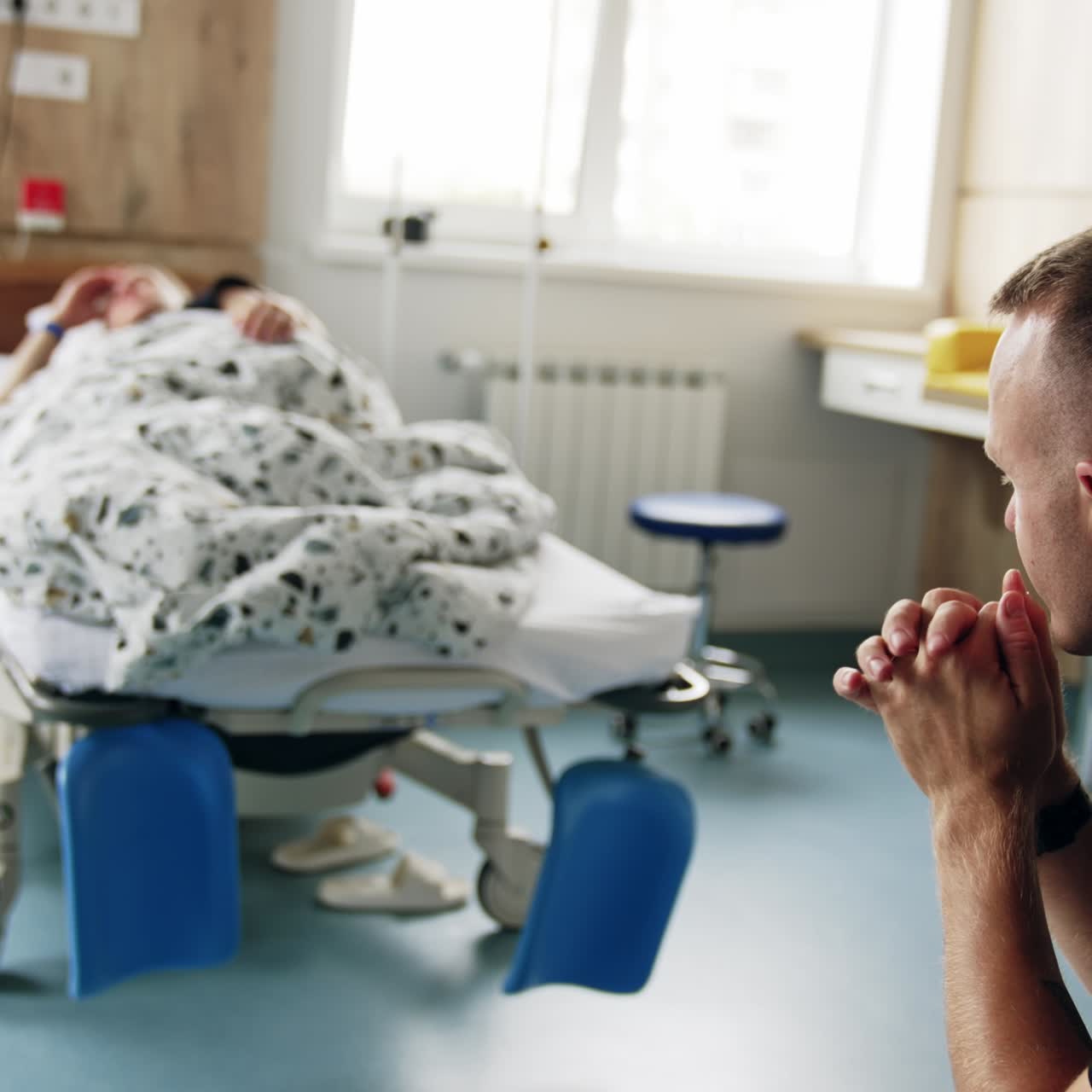 Mid-aged Caucasian man sits in the hospital ward. Pregnant woman lies in bed at backdrop in blur. Husband worries about his wife before labor
