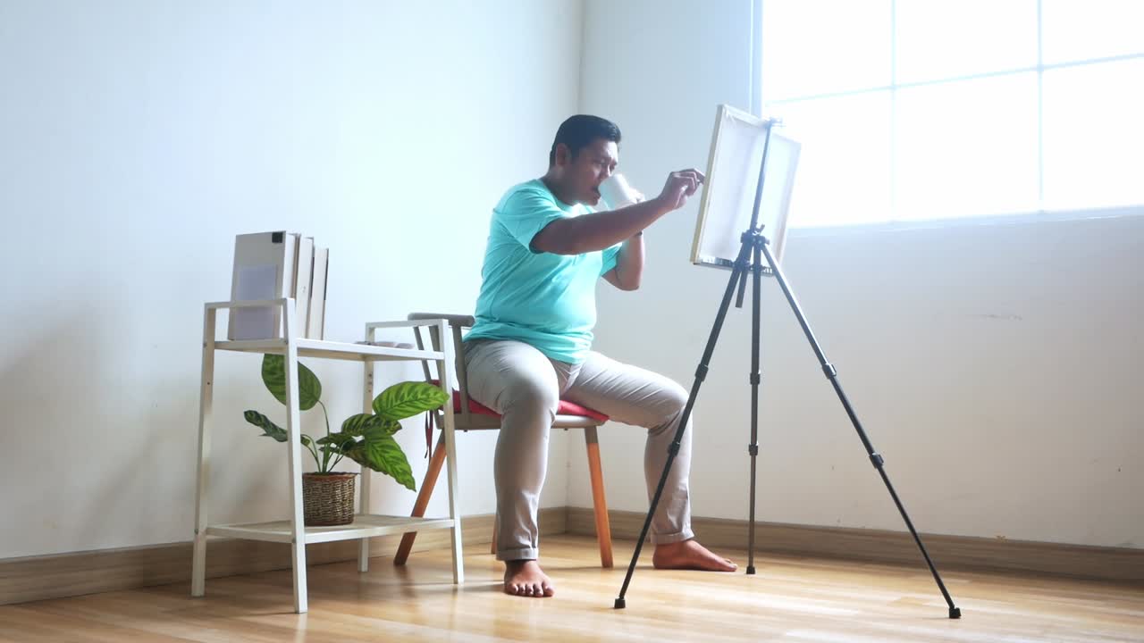 Artist focused on creative process painting canvas at easel holding cup in studio