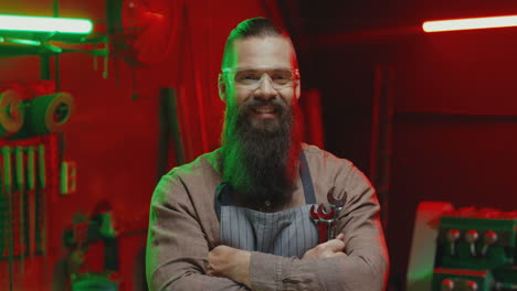 caucasian young man welder with long beard, apron and goggles looking at camera with arms crossed in workshop