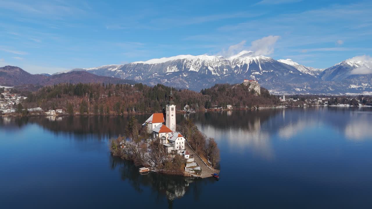 Chiesa sull'Isola di Bled innevata che si riflette nel Lago di Bled con le Alpi sullo sfondo, orbita oraria bassa con drone