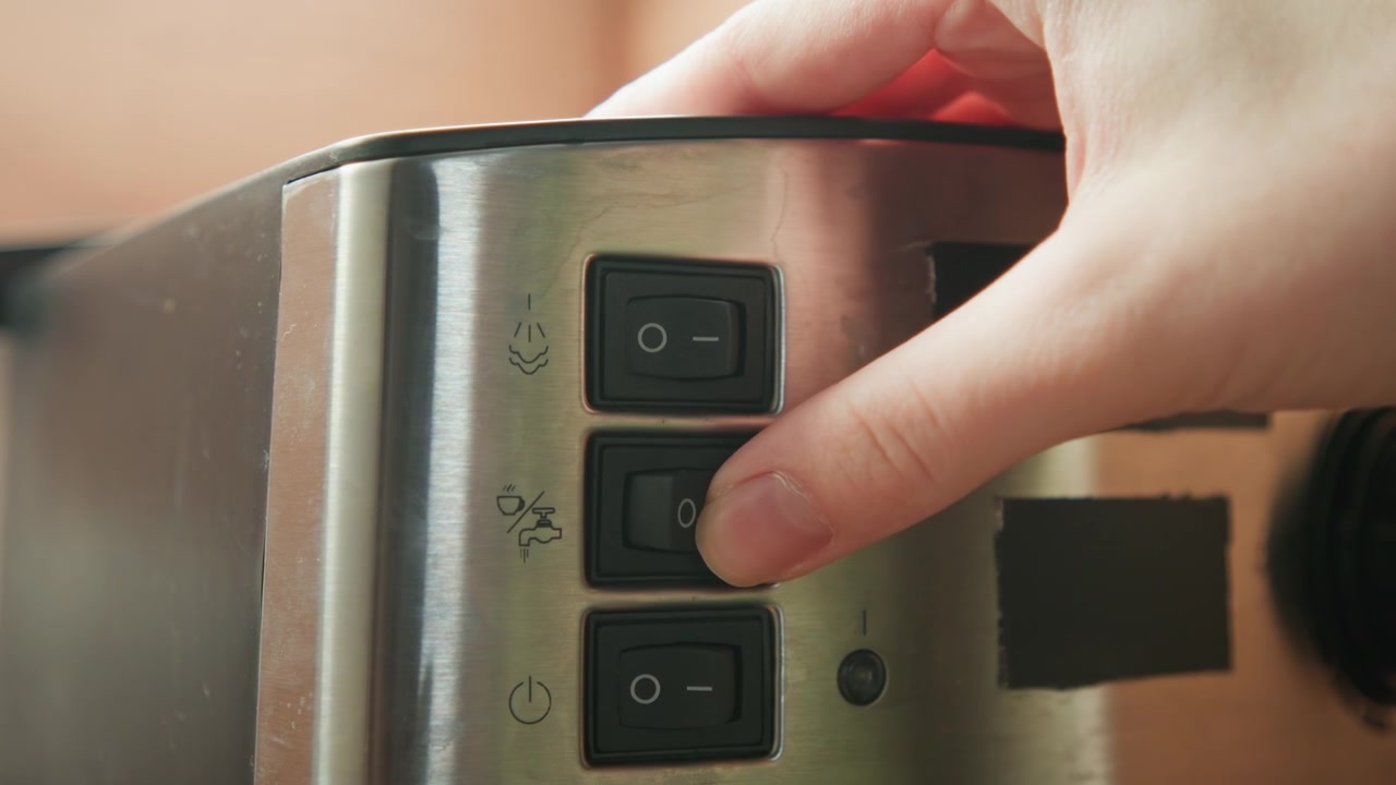 Tight close-up showing stainless steel toaster side with three black rectangular control buttons, center button visibly recessed during interaction; metal casing has black tape strips, smudges, and