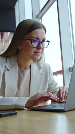 young woman works on her laptop sitting at the desk. lady in glasses looking intently at the screen. workplace at the panoramic window. vertical video