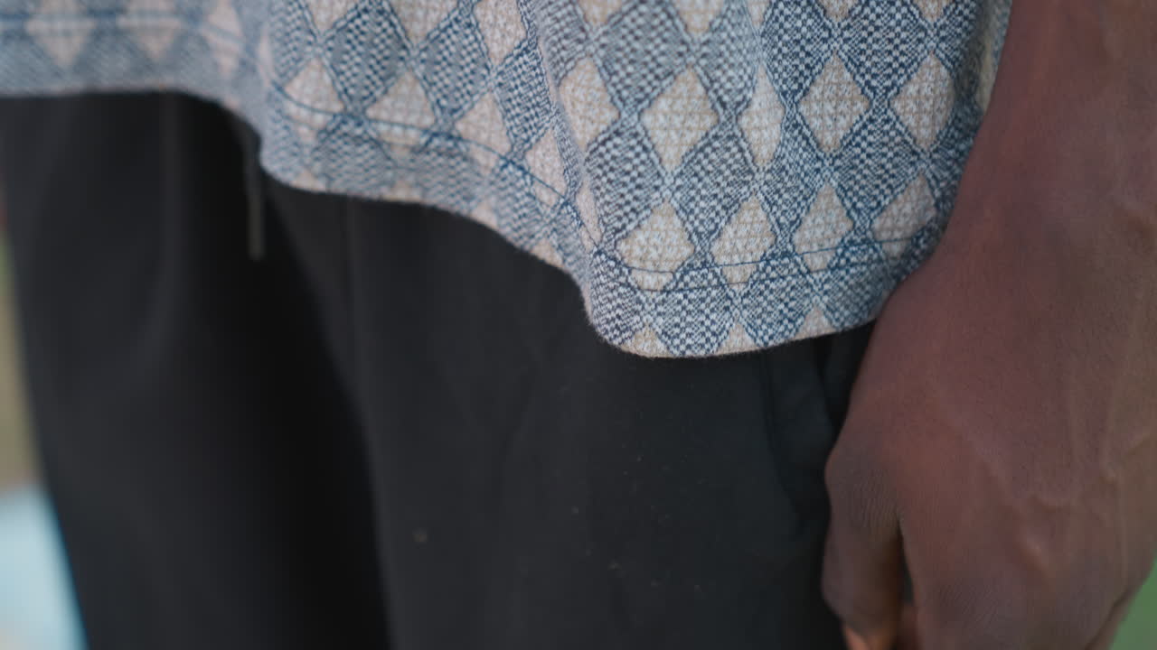 Uomo di colore con la mano in tasca, camicia a fantasia e pantaloncini sportivi neri. Ritratto ravvicinato all'aperto mostra tessuto testurizzato, braccio dalla pelle scura, postura rilassata durante una pausa dall'allenamento, luce solare soffusa e ambiente urbano.