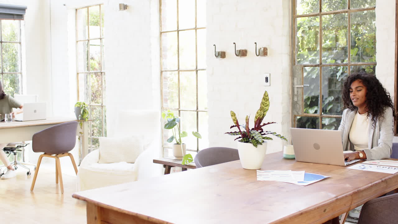 Two multiracial female colleagues collaborating at home office, using laptop and tablet, discussing