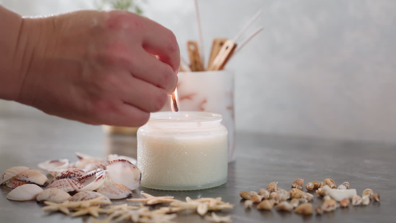 Hand view of person using lit match to light white candle on marble tabletop surrounded by seashells, starfish, and decorative items creating cozy coastal inspired arrangement for calming home decor