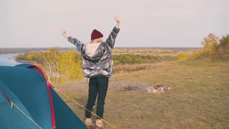 girl tourist stretches at tent and bonfire on river bank