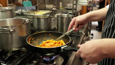 chef cooking and stirring penne pasta in a skillet pan on a stove in the kitchen of a restaurant