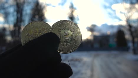 mano sosteniendo dos monedas de bitcoin amarillas doradas y reflectantes durante un día de invierno. las imágenes primero son borrosas y el enfoque cambia y se centra en las monedas de criptomoneda. cielo azul y nublado visto.