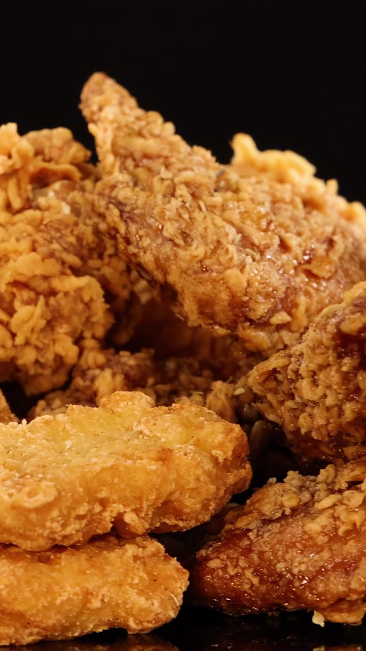 Golden fried chicken wings, drumsticks, and nuggets rotate on a reflective surface under warm studio lighting, revealing crunchy textures against a black background