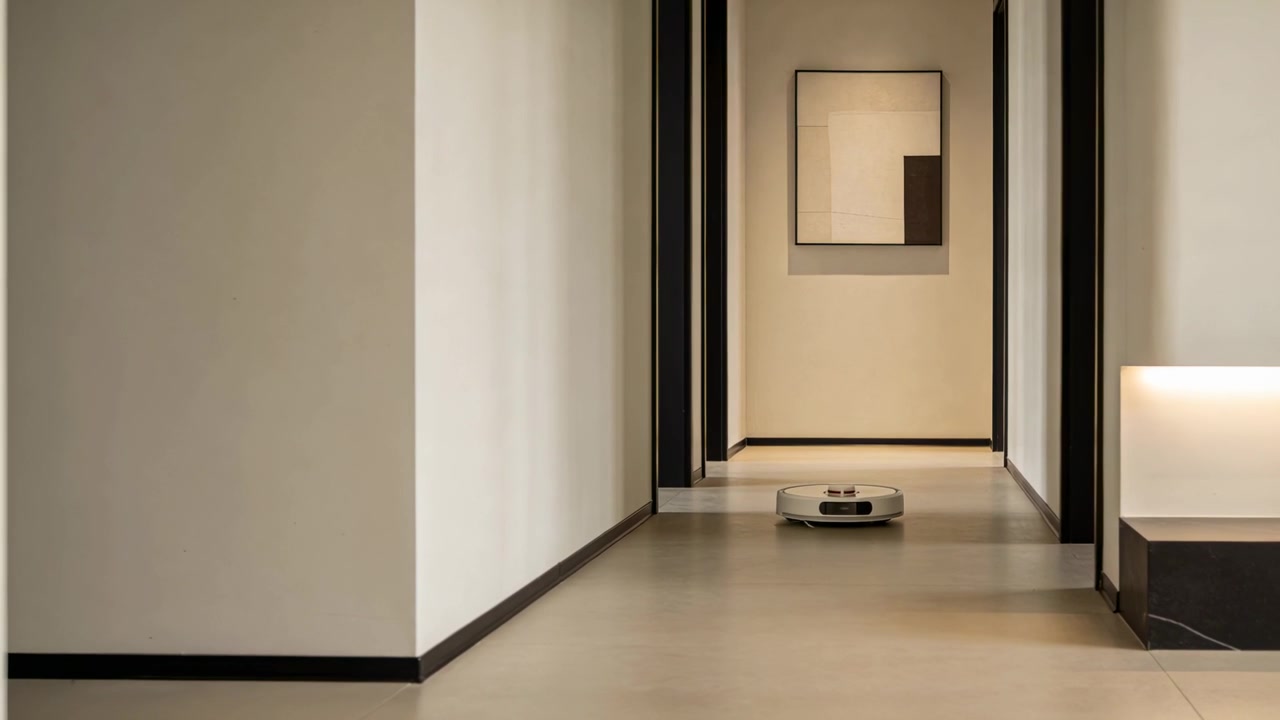 Aspirapolvere robot che parte vicino a un'opera d'arte incorniciata, percorrendo il corridoio di casa verso la telecamera in ciclo di pulizia.