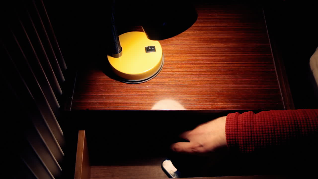 밤에 한 남자의 손이 작은 램프의 빛을 켜고 스탠드의 서랍에서 스마트폰을 뽑고 있습니다.