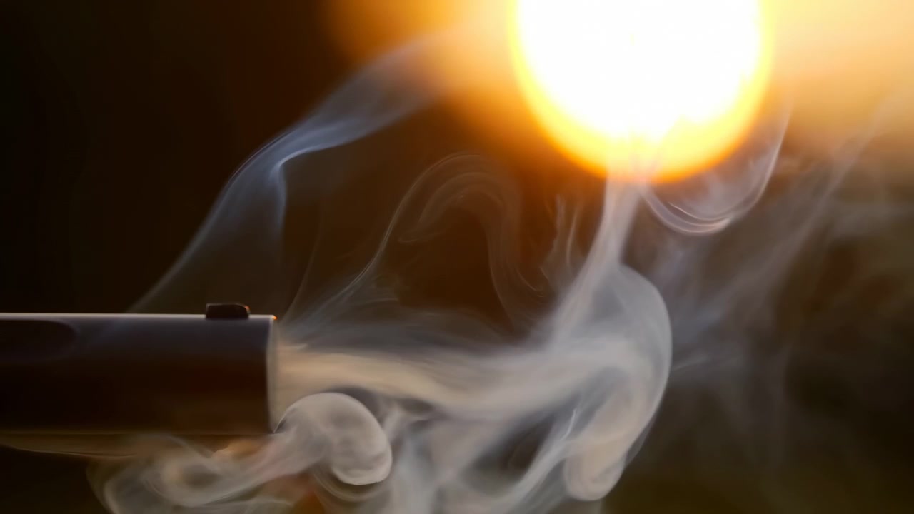 Side view of handgun discharge showing bright orange yellow muzzle flash and thick white gray smoke curling diagonally through dark low key lighting, creating tense dramatic contrast