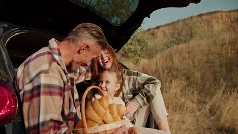 A-man-with-gray-brunette-hair-communicates-with-his-happy-wife-in-a-green-checkered-shirt-and-their-little-daughter-a-blonde-girl-while-they-sit-in-an-open-trunk-during-their-picnic-outside-the-city-in-the-summer