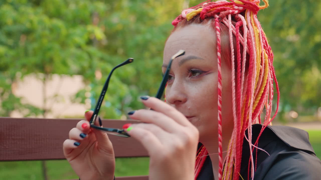 Mujer ajustándose las gafas, rastas de colores en un parque, retrato de una mujer con rastas y estado de ánimo reflexivo, escena al aire libre con una mujer con rastas vibrantes y expresión contemplativa