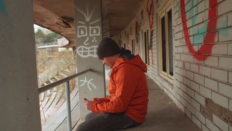 man in orange jacket and beanie sits scrolling on phone in old abandoned stadium