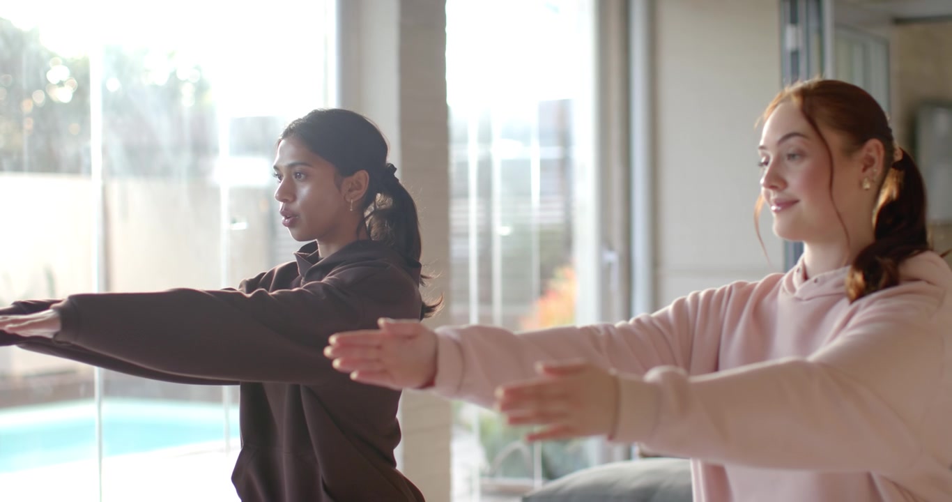 Diverse women in hoodies after left cue, doing squats and arm reaches for workout by pool