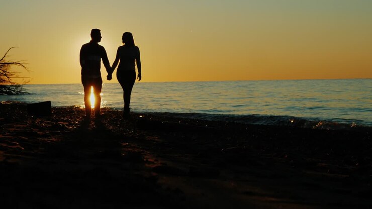 Silhouettes d'un couple amoureux marchant au coucher du soleil au bord de la mer ou d'un lac. Magnifique reflet du soleil. Homme asiatique et femme caucasienne.