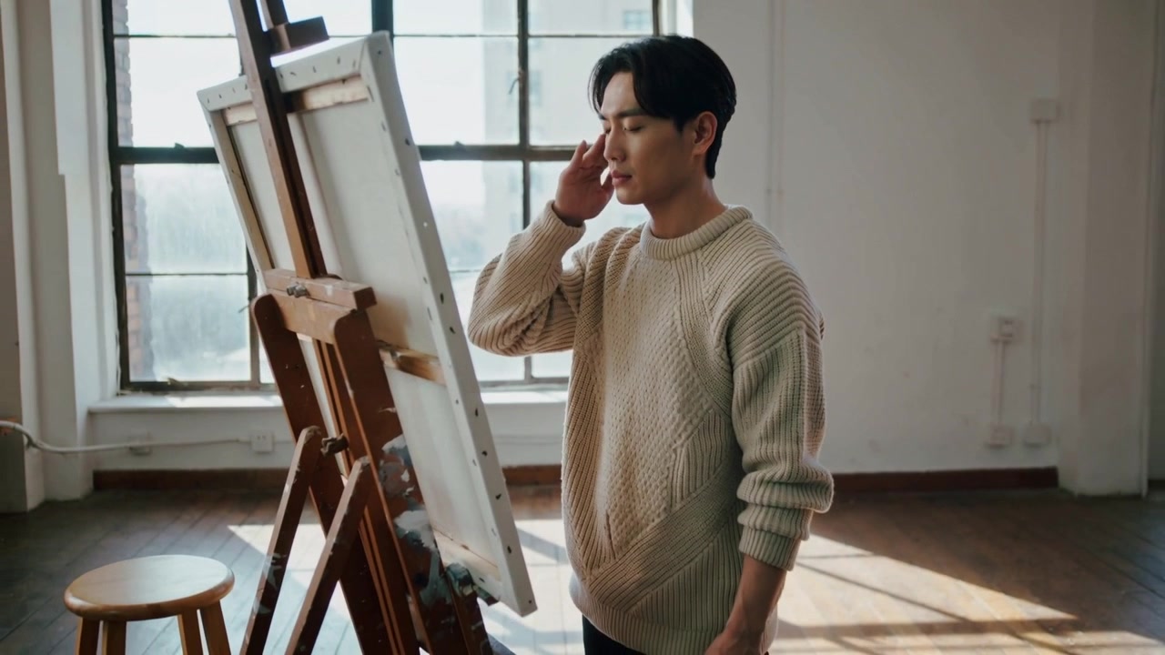 햇살 가득한 로프트에서 관자놀이를 만지며 캔버스를 평가하고 이젤 앞에서 그림을 구상하는 스웨터 차림의 예술가