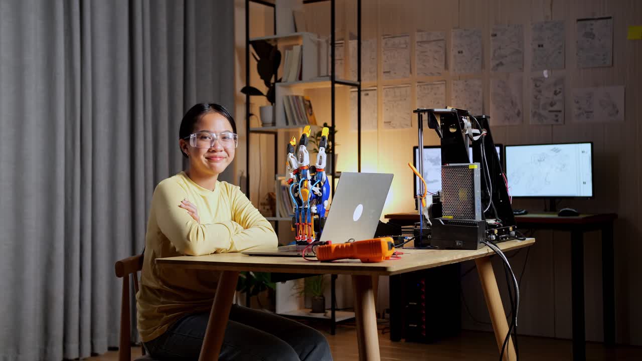 집에서 사이보그 손에 대해 작업하는 동안 노트북을 들고 있는 십대 아시아 소녀가 미소 짓고 카메라에 팔을 고 있습니다.